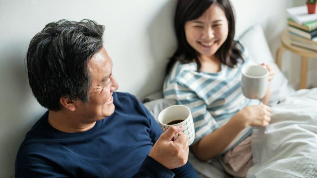 A couple smiles and talks in bed while enjoying mugs of coffee or tea.