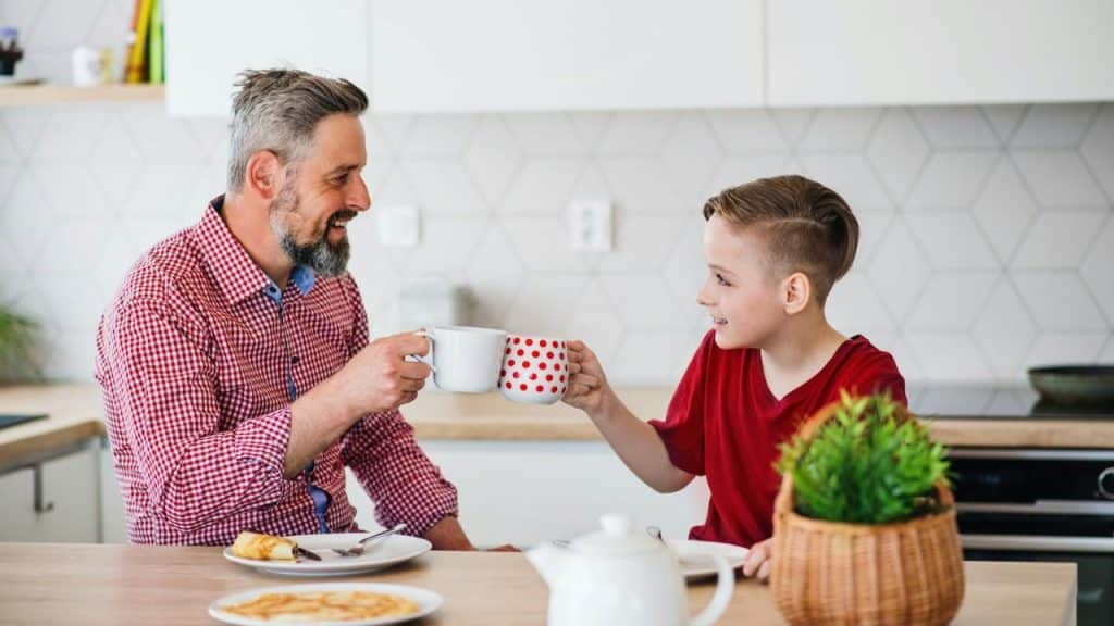 A smiling father and son in a kitchen cheers mugs over a table with breakfast.