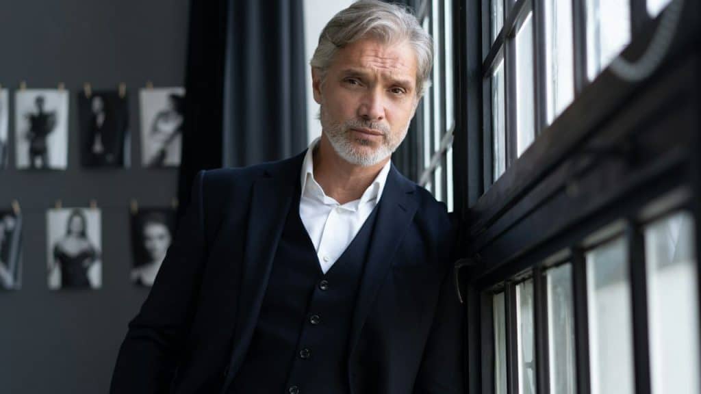 A serious, silver-haired man in a dark suit leans against a window.