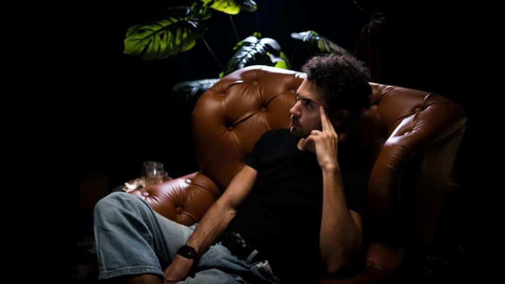 A man in a black shirt and jeans sits thoughtfully in a leather armchair.