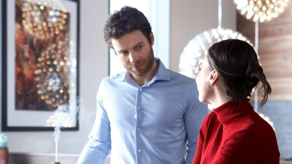 A man in a blue shirt attentively listens to a woman in a red turtleneck indoors.