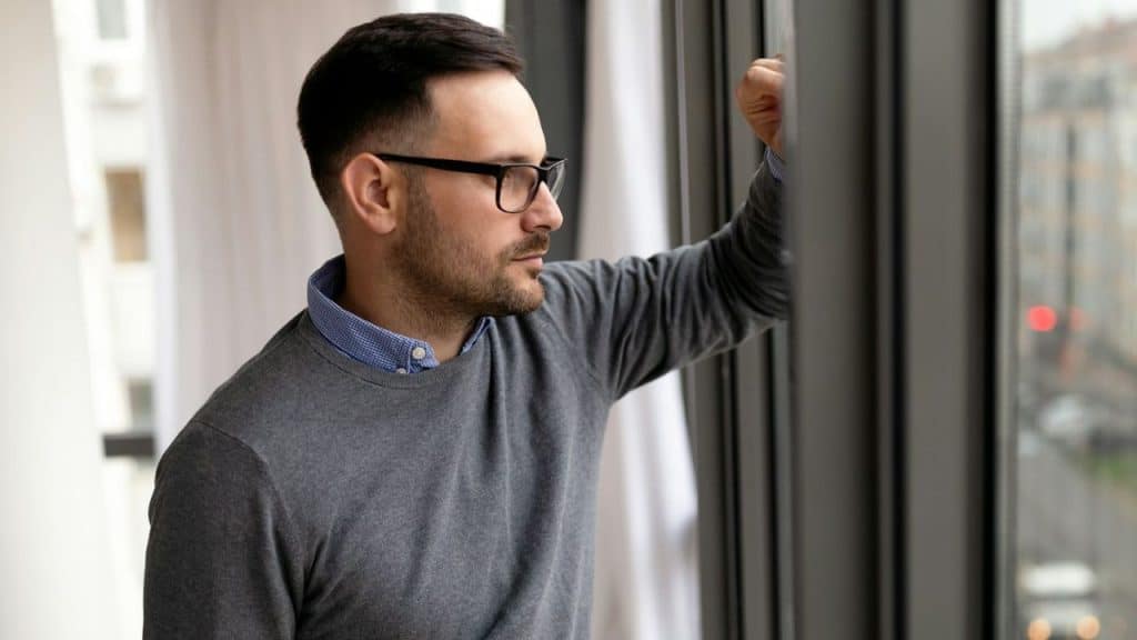 A thoughtful man in glasses and a grey sweater looks out a window.