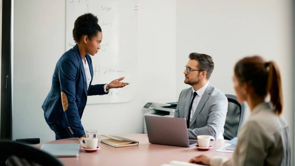 A professional woman gestures while speaking to a man and another person at a meeting table.