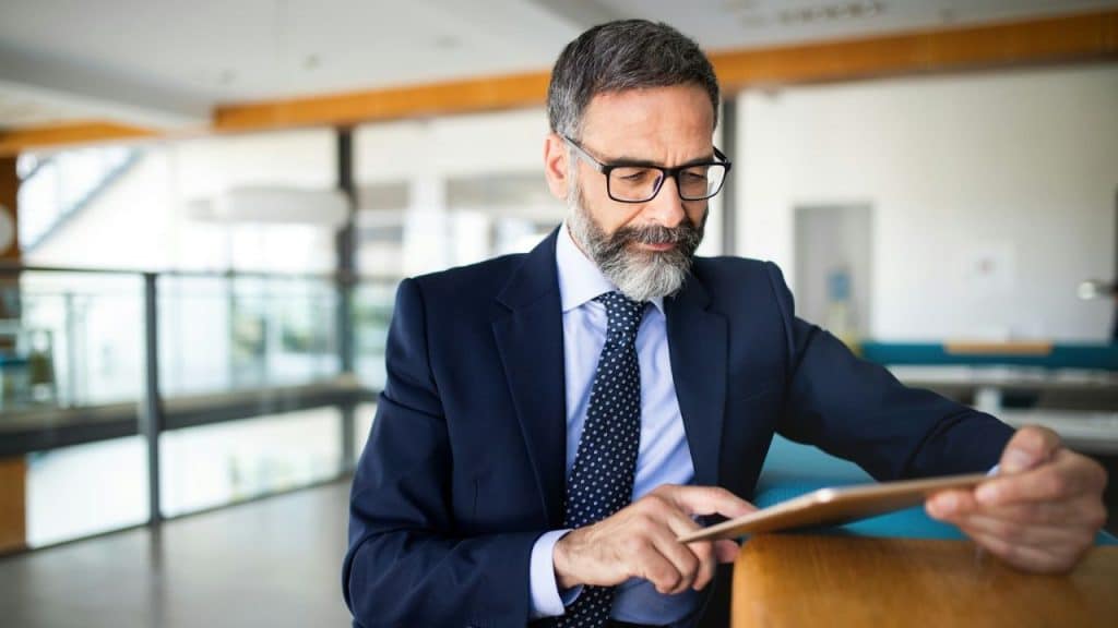 A distinguished, bearded businessman in a suit uses a tablet in a bright office.