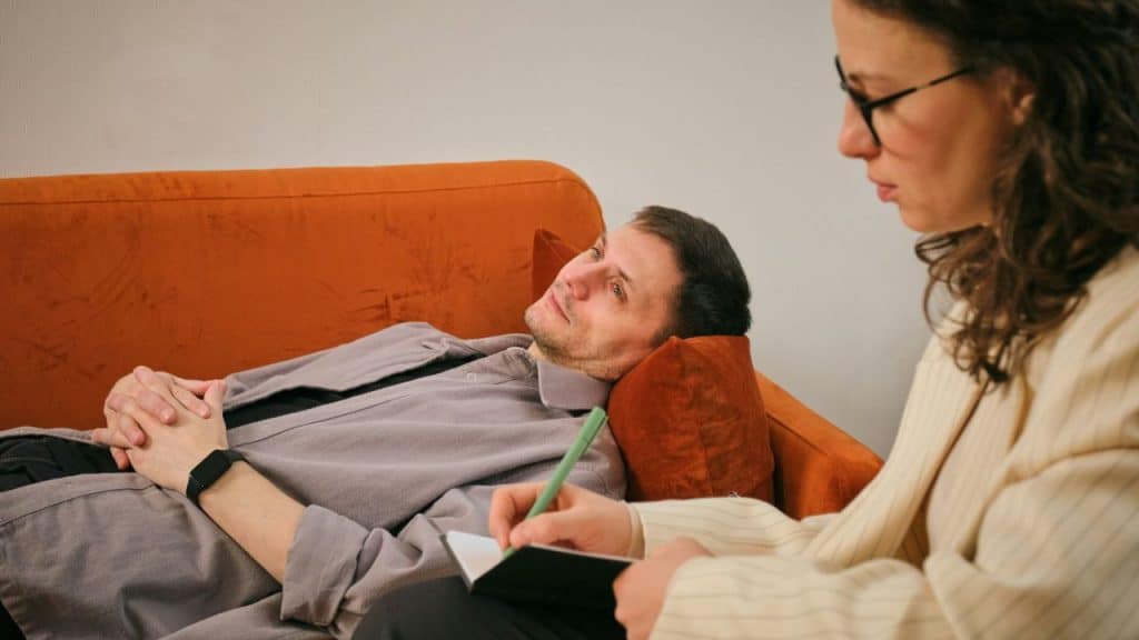 A man lies on an orange couch talking to a therapist who is writing on a notepad.