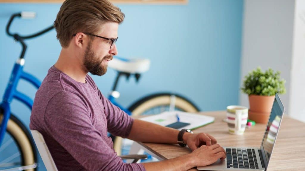 A bearded man in glasses works on a laptop at a desk with a blue bicycle behind him.