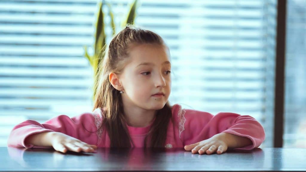 A young girl in a pink sweater rests her hands on a dark table.