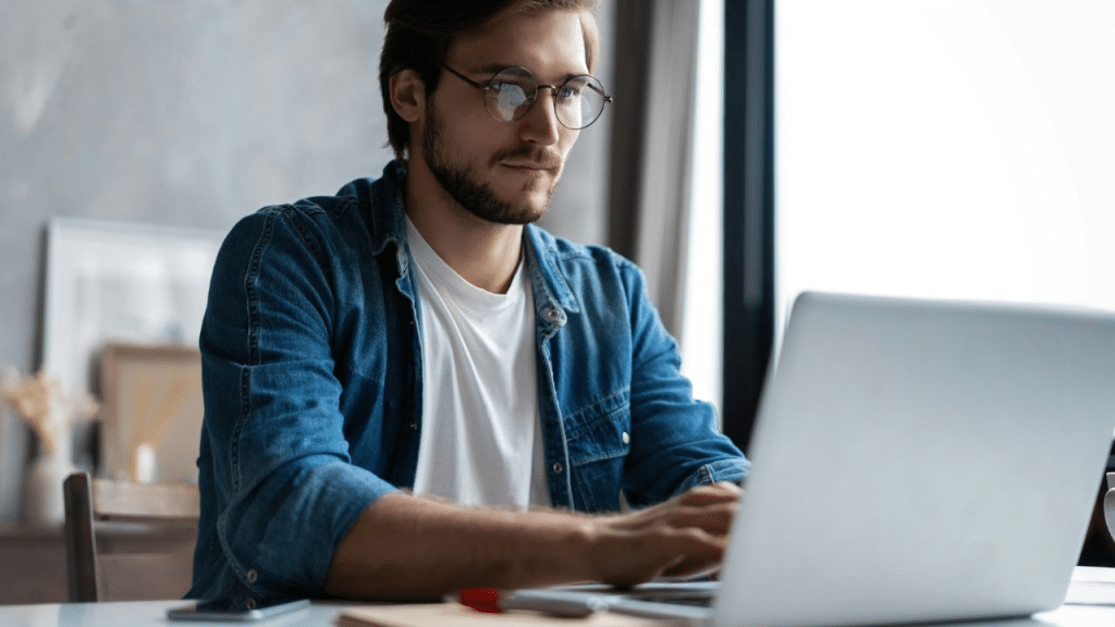 A handsome, bearded man in glasses works seriously on his laptop at home.