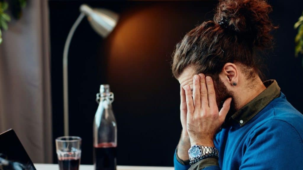 A man with his hair in a bun holds his hands to his face, looking stressed.