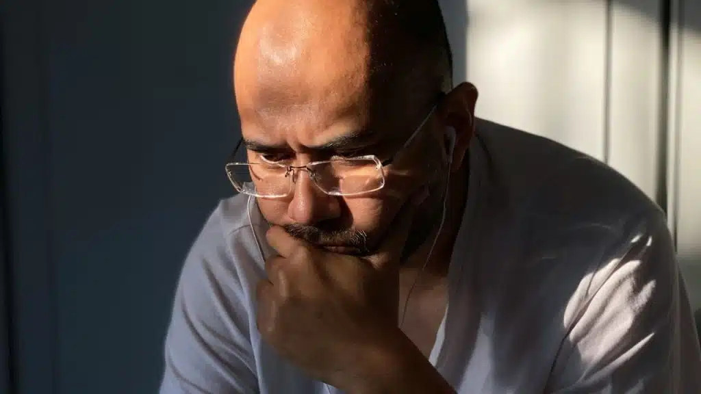 A balding man in glasses and earbuds looks down thoughtfully in shadow.