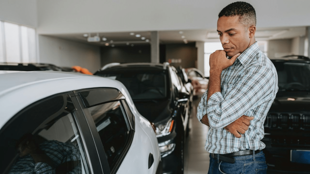 A serious, thoughtful man in a plaid shirt examines a car at a dealership.