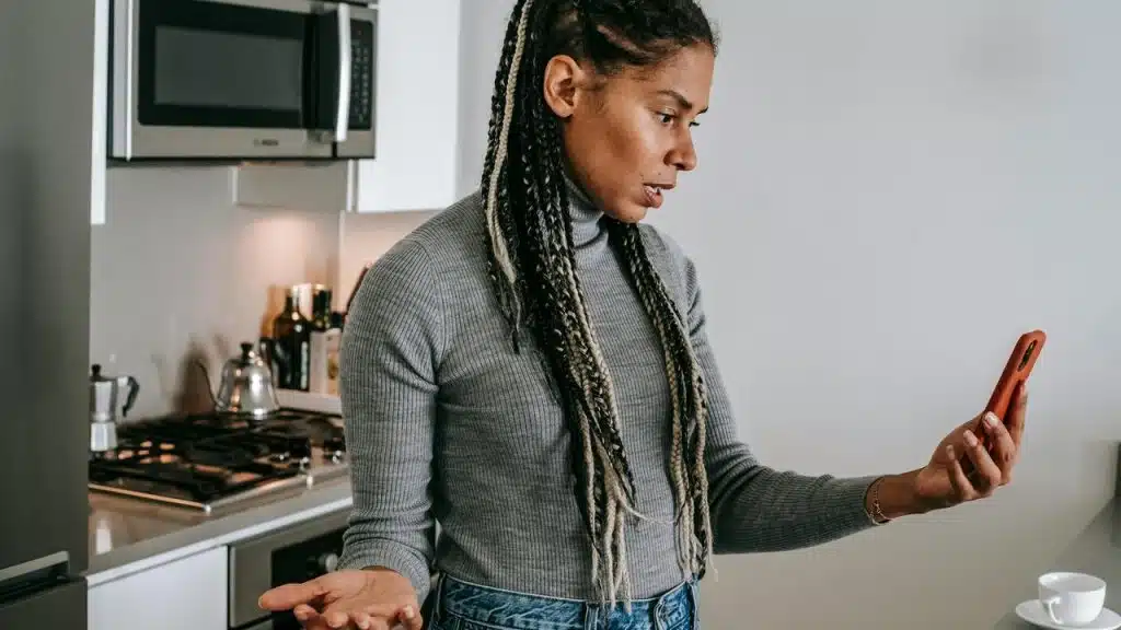 A woman standing in a kitchen holding her phone and looking frustrated.