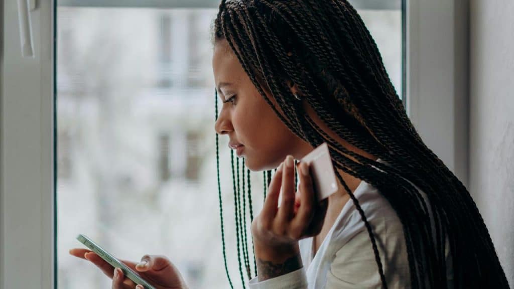 A woman holding a credit card and looking at her phone near a window.