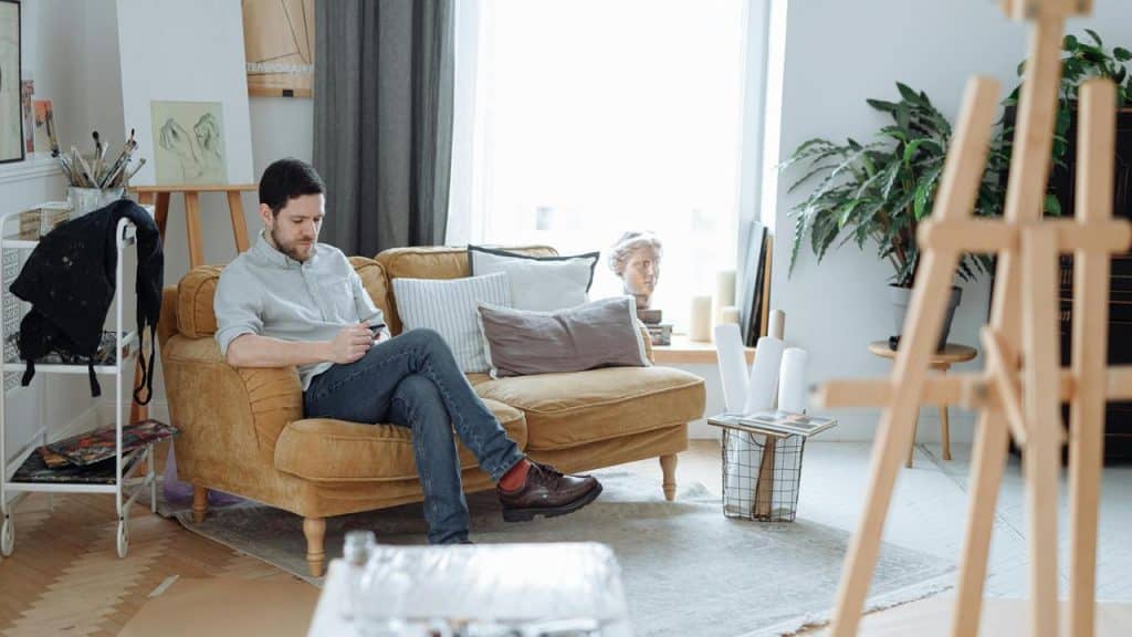 A man sitting on a couch in a bright living room looking at his phone.