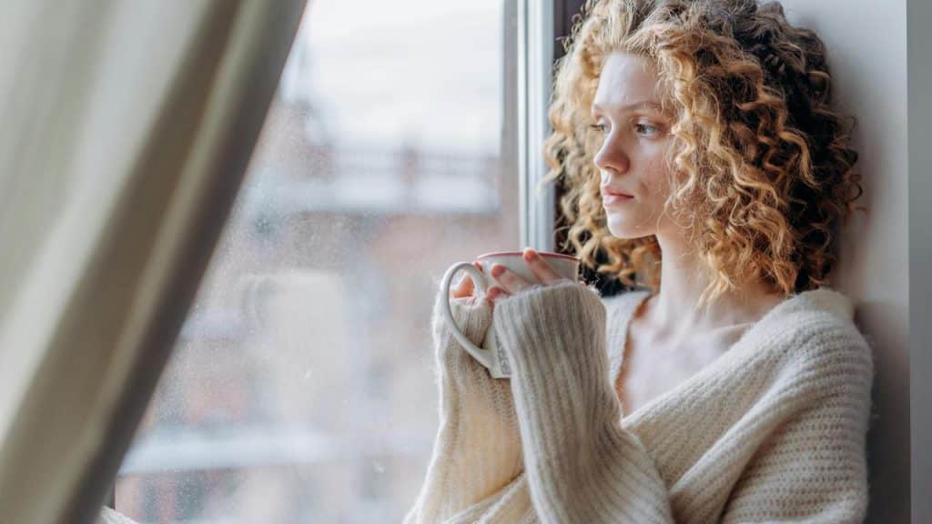 A woman with curly hair sitting by a window holding a mug and looking outside.