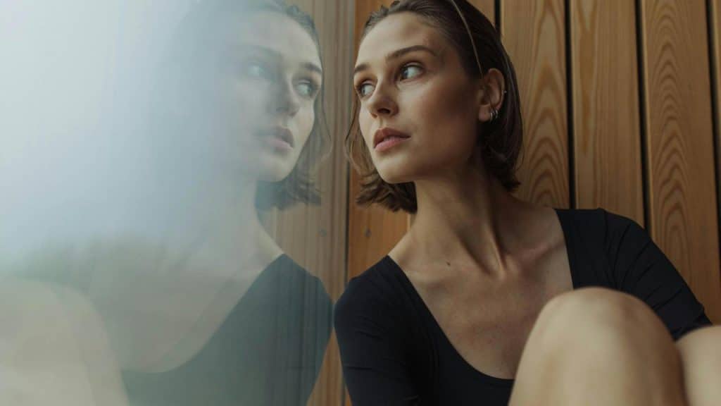A woman sitting by a window looking thoughtful with her reflection visible in the glass.