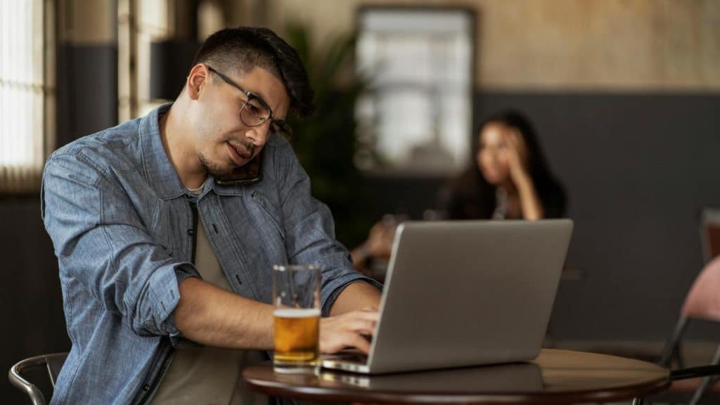 A man talking on the phone while working on a laptop in a café.