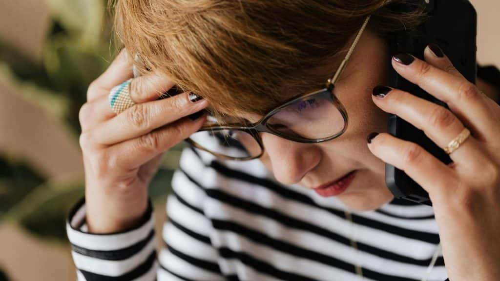 A woman wearing glasses talking on the phone with a concerned expression.