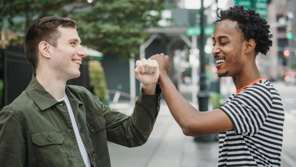 A pair of friends smiling and greeting each other with a fist bump on a city street.