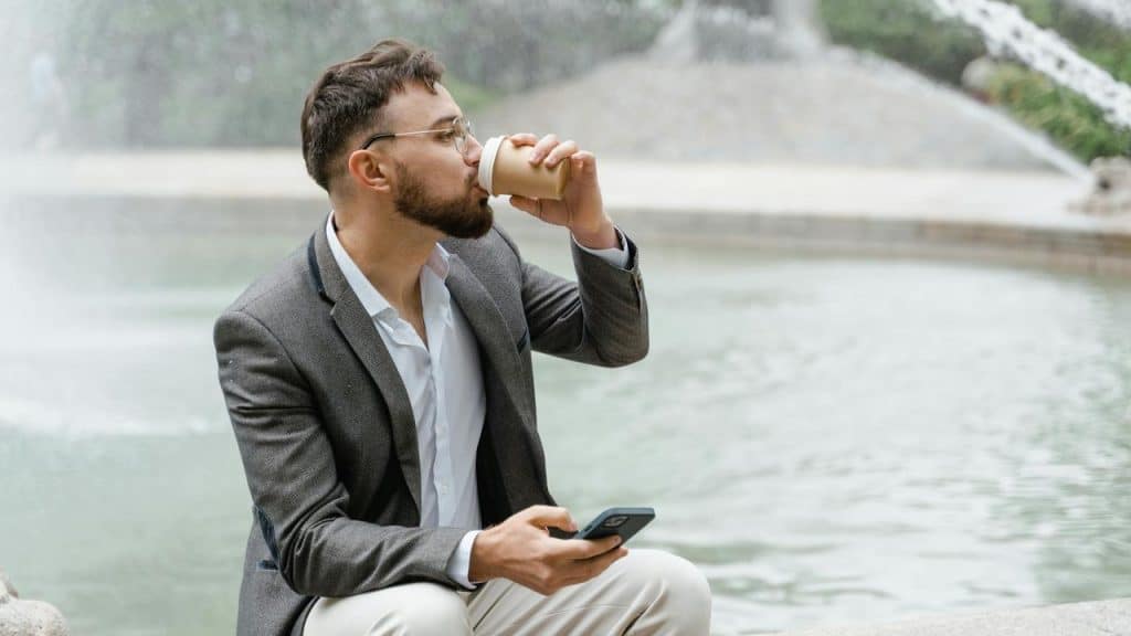 A man in a suit sitting by a fountain drinking from a paper cup and holding a phone.