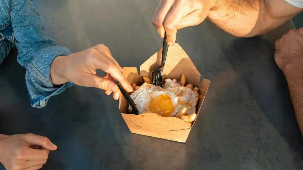 A pair of hands using forks to share a meal with fried eggs in a takeout box.