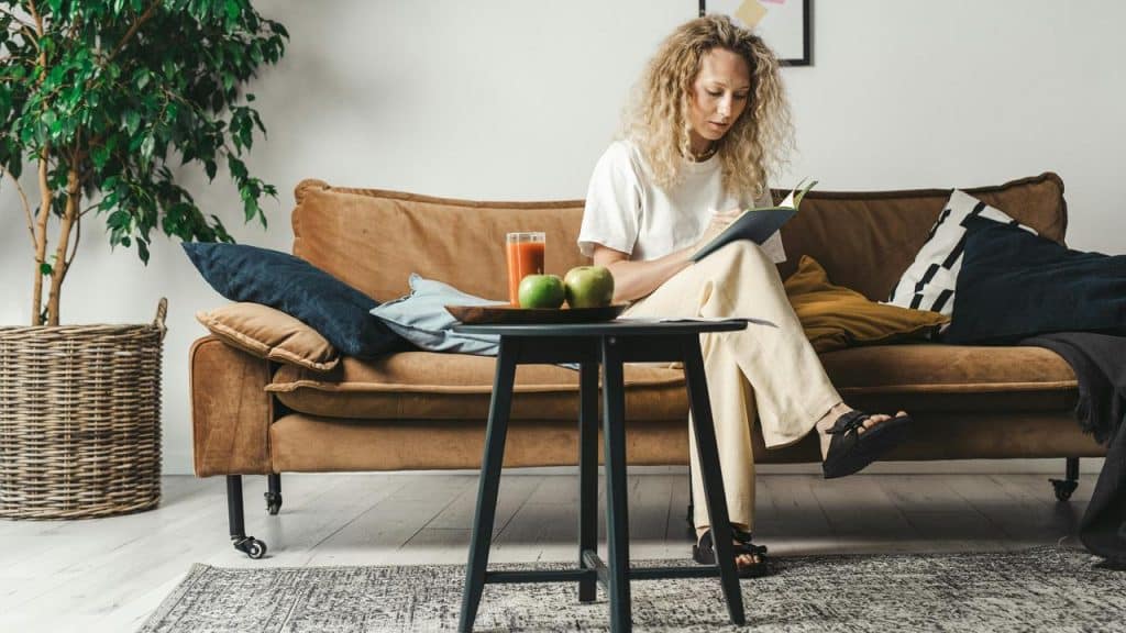 A woman sitting on a couch writing in a notebook with a drink and apples on the table.