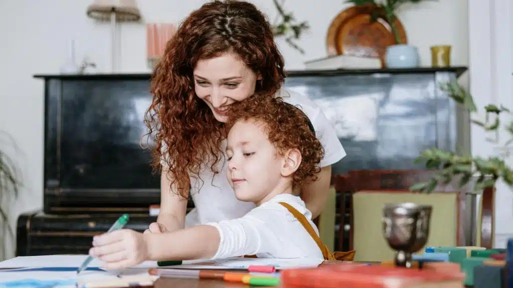 A mother smiling while helping her child draw at a table.