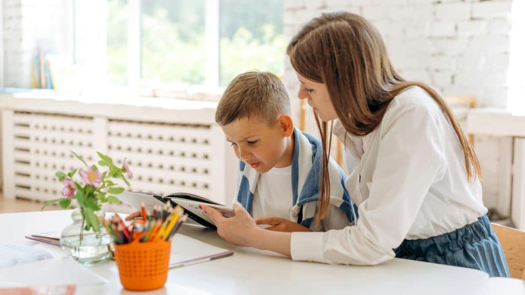 A mother helping her son read a book at a bright table.