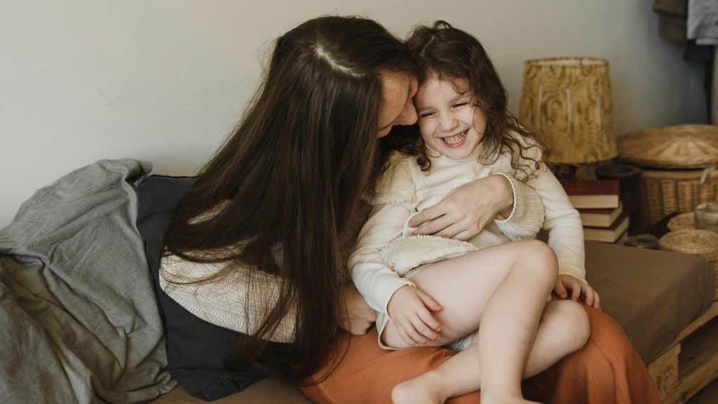A mother laughing while hugging her daughter on the couch.