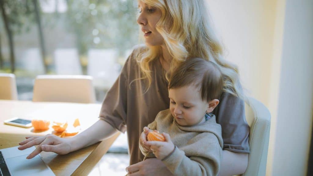 A mother working on a laptop while holding her baby eating an orange.