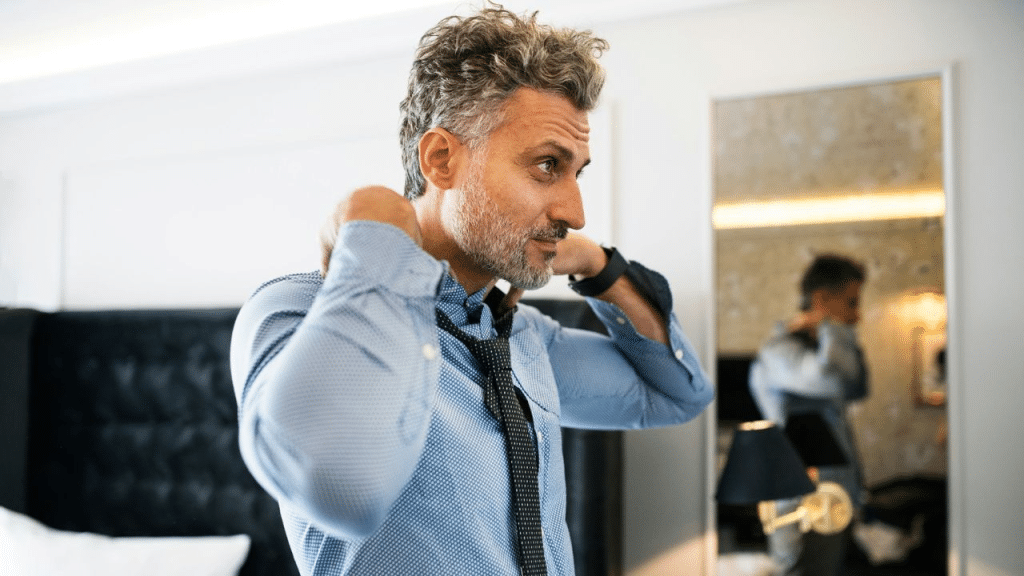 A handsome middle-aged man adjusts his tie in front of a mirror in a bedroom.