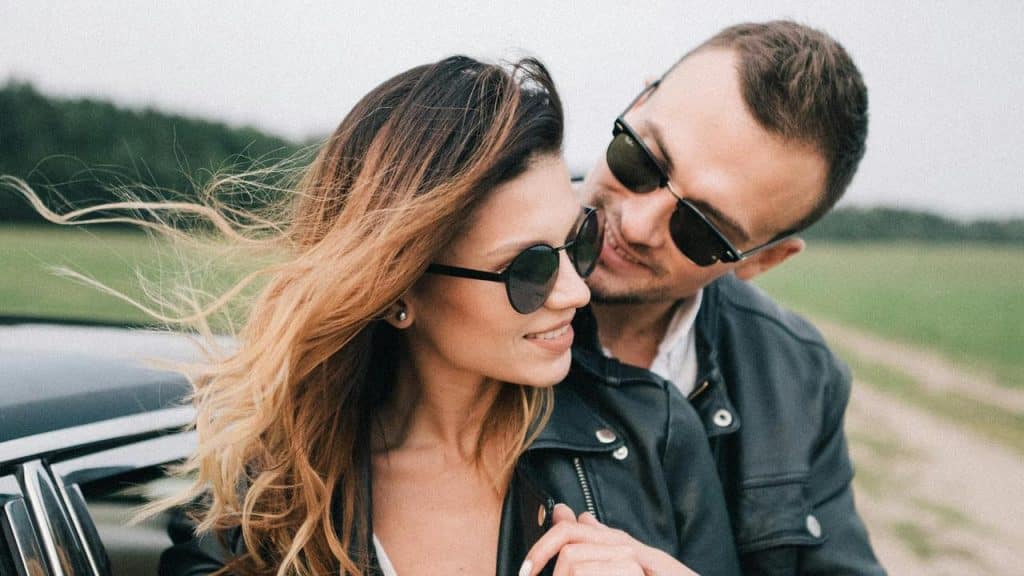 A couple wearing sunglasses smiling at each other beside a car in the countryside.
