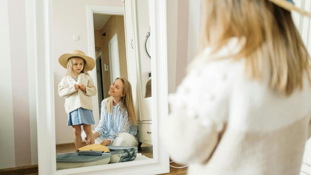 A mother smiling at her daughter trying on a hat in front of a mirror.