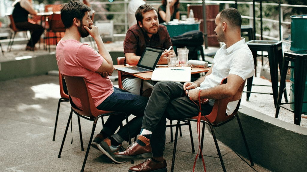 Three men sit at a small outdoor table, engaged in a serious conversation.