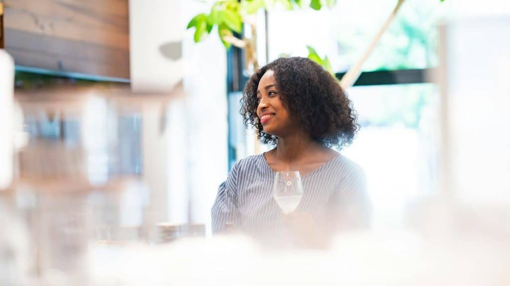 A woman smiling while holding a glass of white wine in a bright café.