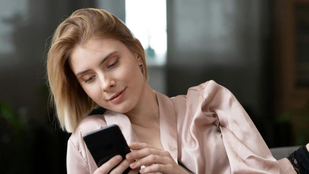 A woman lying on a bed smiling while using her smartphone.