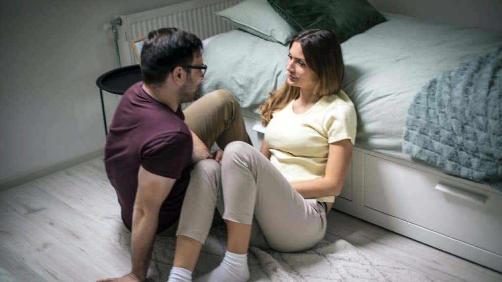 A man and woman sitting on the floor by a bed having a serious conversation.