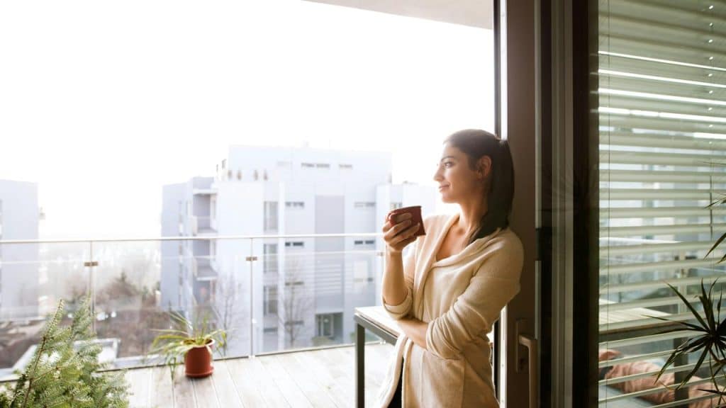 A woman standing by a balcony door holding a cup and looking outside.