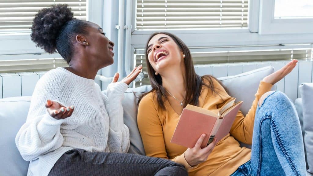 A woman laughing with a friend while holding a book on a couch.