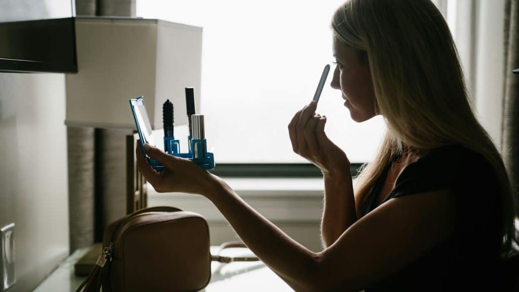 A woman sitting by a window applying makeup with a mirror.