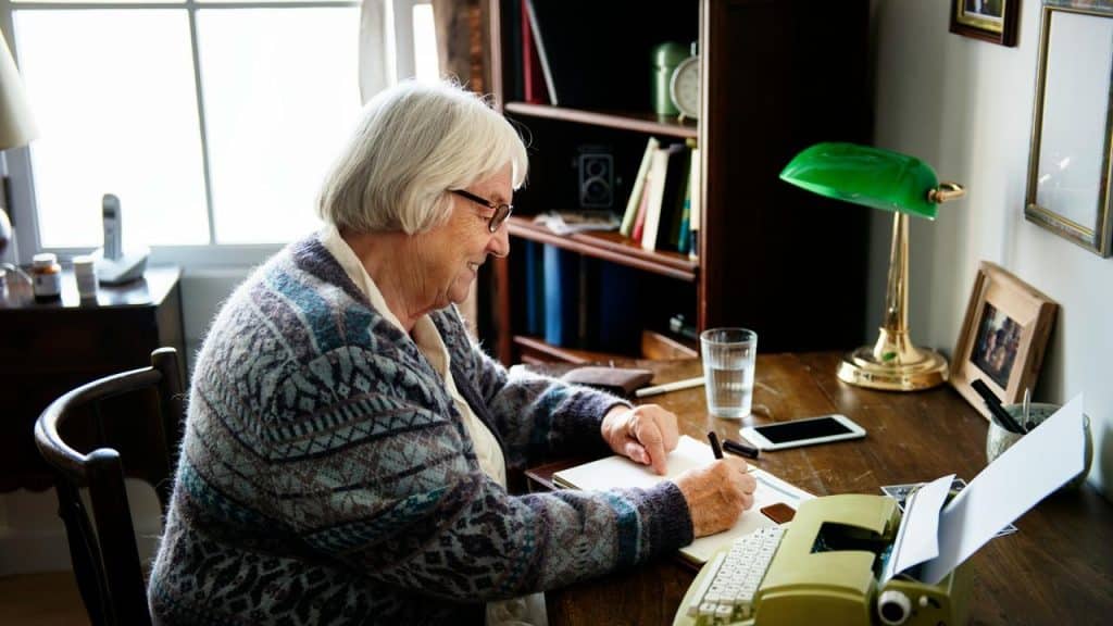 An elderly woman writing at a wooden desk with a typewriter and a green lamp.