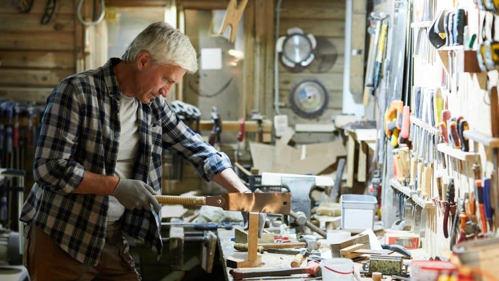 An older man working with a hand saw in a cluttered woodworking shop.