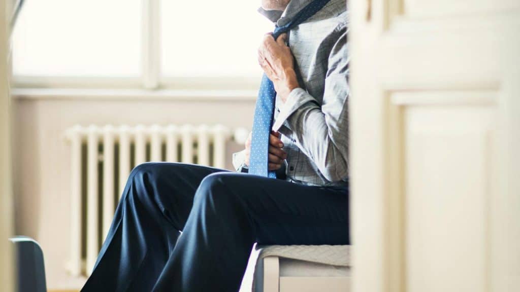 An older man sitting on a bed and adjusting his blue tie.