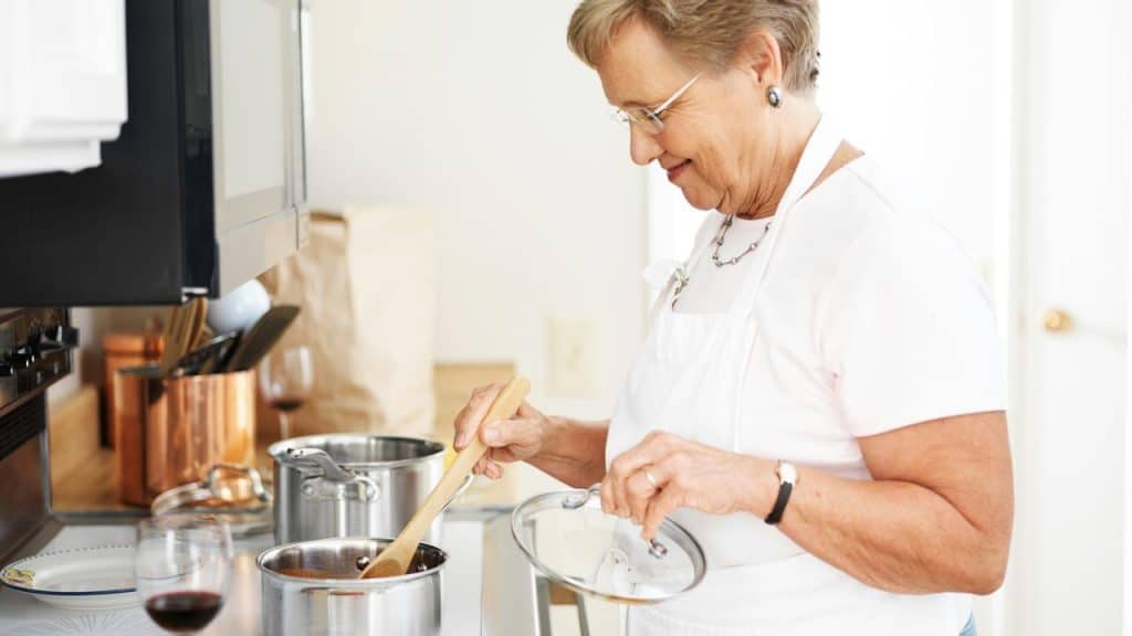 An elderly woman wearing an apron stirring food in a pot on the stove.