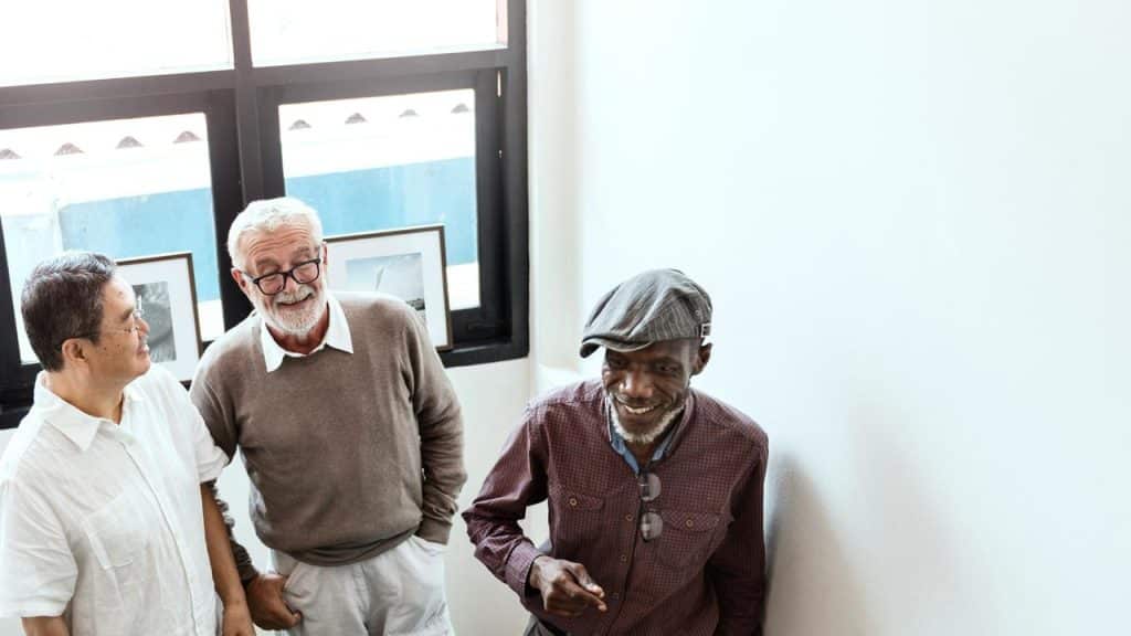A group of three older men standing indoors and smiling while talking.