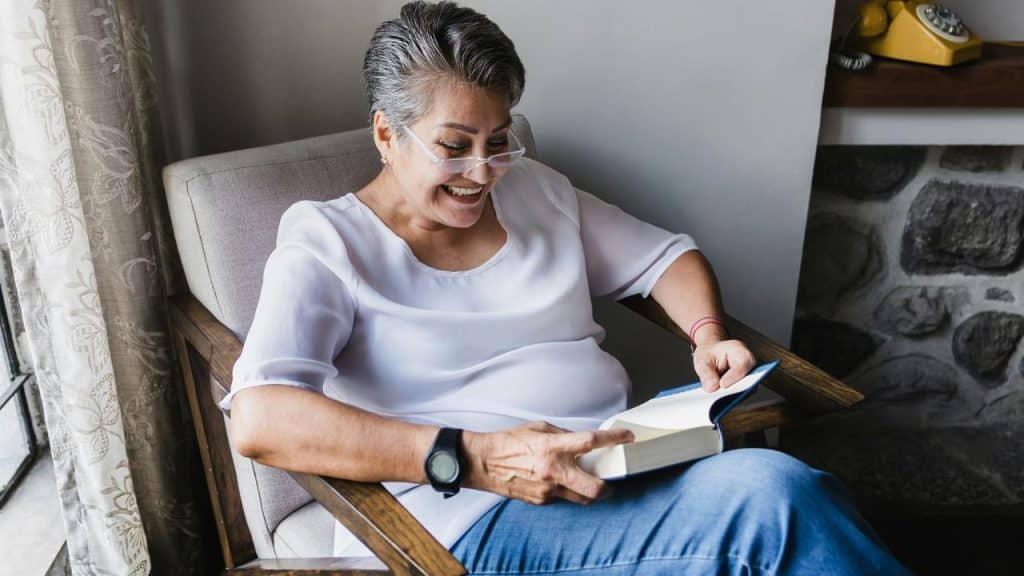 An older woman sitting by a window smiling while reading a book.