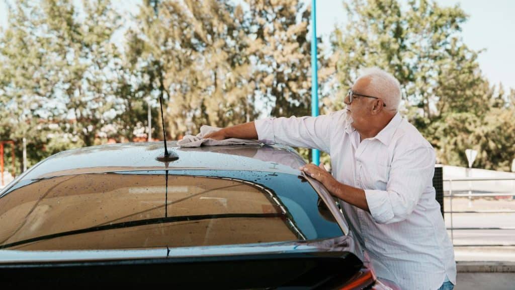 An older man cleaning the roof of his car with a cloth outdoors.