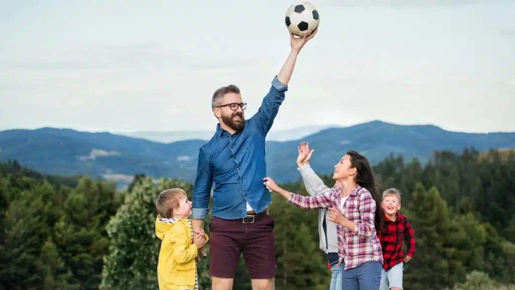 A bearded man holds a soccer ball high above a group of three children on a grassy hill with forested mountains in the background.