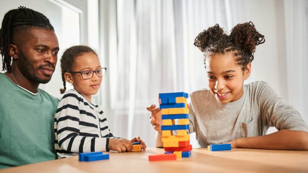 Three people, an adult male and two girls, are sitting at a table playing a colorful block stacking game indoors.