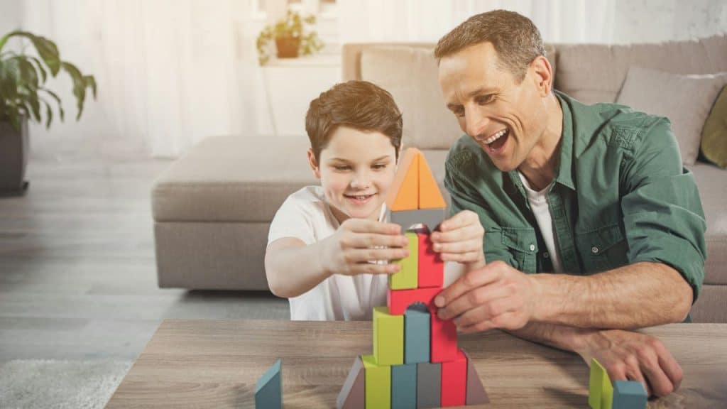 A smiling man and boy are leaning over a wooden table building a colorful block structure together.